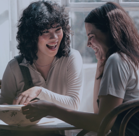 Kaksi ruskeatukkaista naista katsoo lehteä ja hymyilee samalla toisille hampaat näkyen